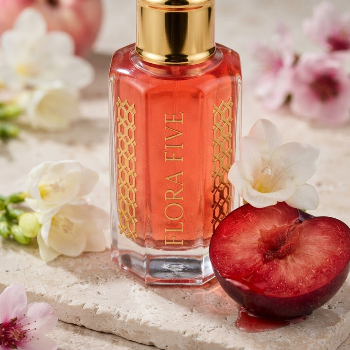 Victoria's Secret Pure Seduction Inspired Attar by Flora Five — close-up of a gold-capped bottle of coral-red oil on travertine with a blood plum, freesia and pink blossoms – 3, 6 and 12 ml