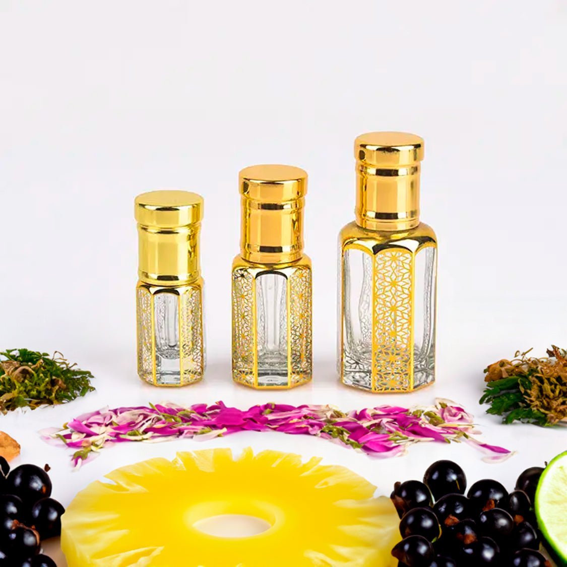 Flora Five Aventus Creed inspired fragrance attar bottles displayed in three sizes with intricate golden designs. Surrounded by natural ingredients including pineapple slices, black currants, floral petals, and greenery, this long-lasting and charming attar embodies luxury and elegance. Perfect for those seeking a captivating and enduring scent.