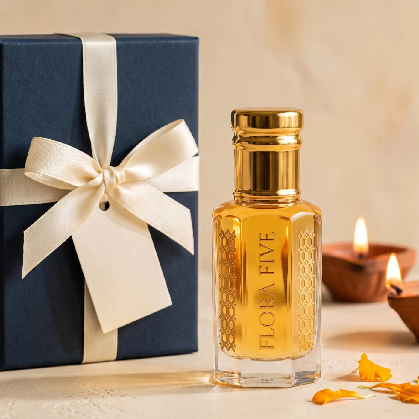 Diwali Gift Box by Flora Five — navy box tied with an ivory satin ribbon and tag beside a golden attar bottle, lit diyas and marigold petals